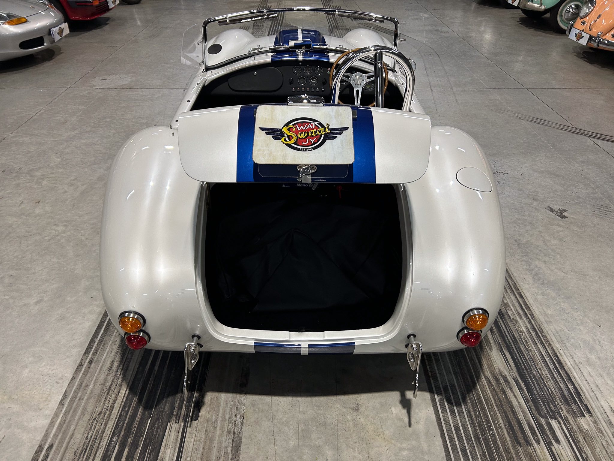 Rear view of a silver vintage race car with a blue racing stripe in a showroom, open cockpit and visible steering wheel panel