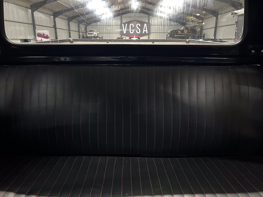 Black leather rear bench seat with vertical orange stitching; visible garage with cars through the rear window.