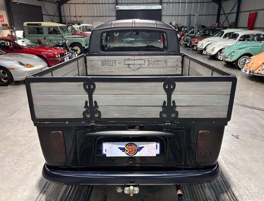 Rear view of a black vintage pickup with a wooden bed and decorative metal accents in a classic car showroom, Harley‑Davidson branding on the tailgate wall panel behind the cab.