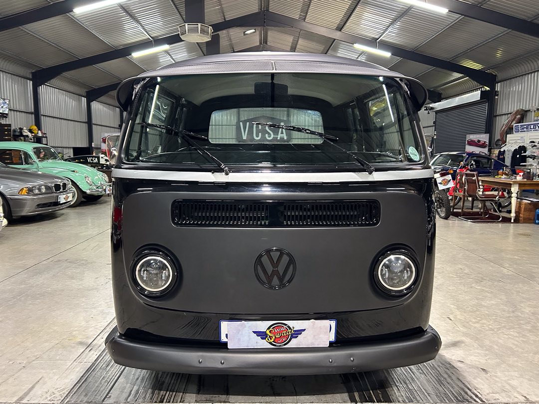 Front view of a black Volkswagen camper van inside a showroom, with other classic cars in the background.