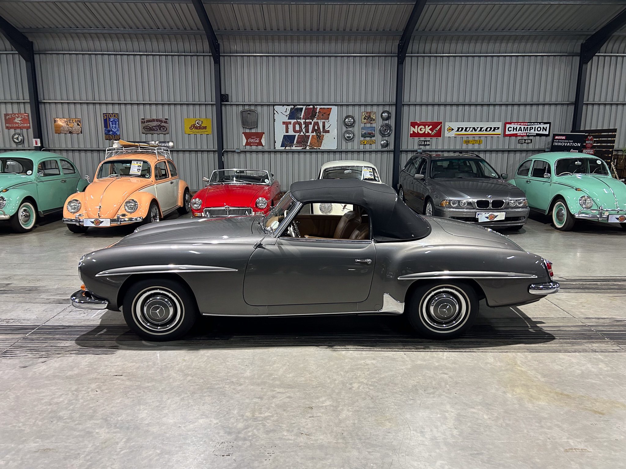 Line of classic cars in a showroom, teal to orange, with vintage posters on a corrugated metal wall behind them, row of vehicles on display.]?