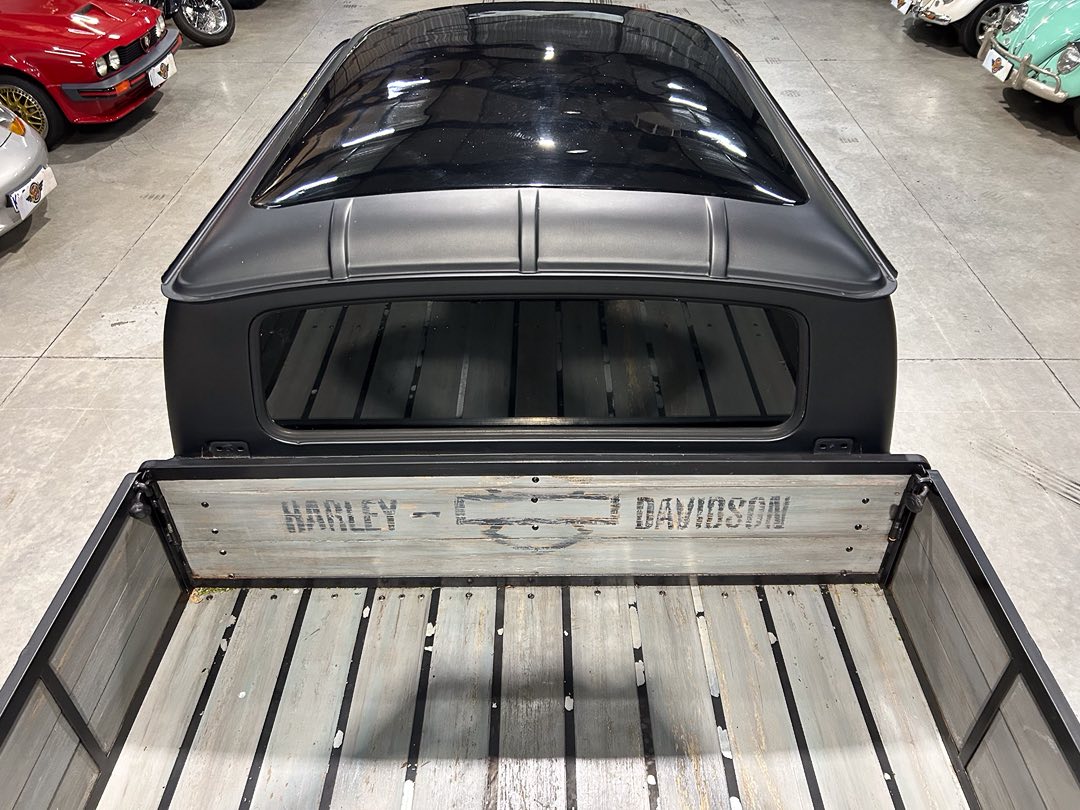 Matte black pickup with a wooden bed and Harley-Davidson logo on the tailgate in a showroom with classic cars nearby.