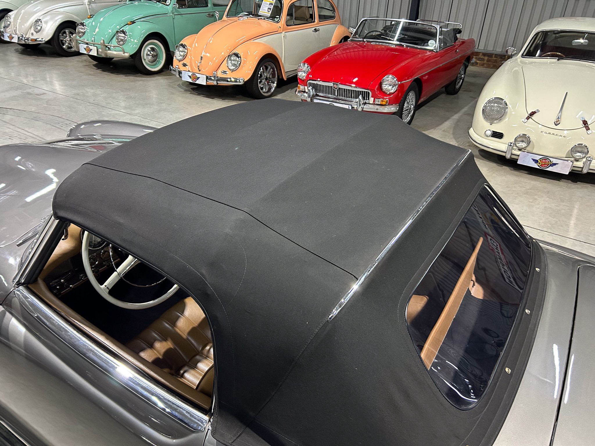 Interior of a vintage convertible with a black fabric roof and tan leather seats, parked among a row of classic cars in a showroom.