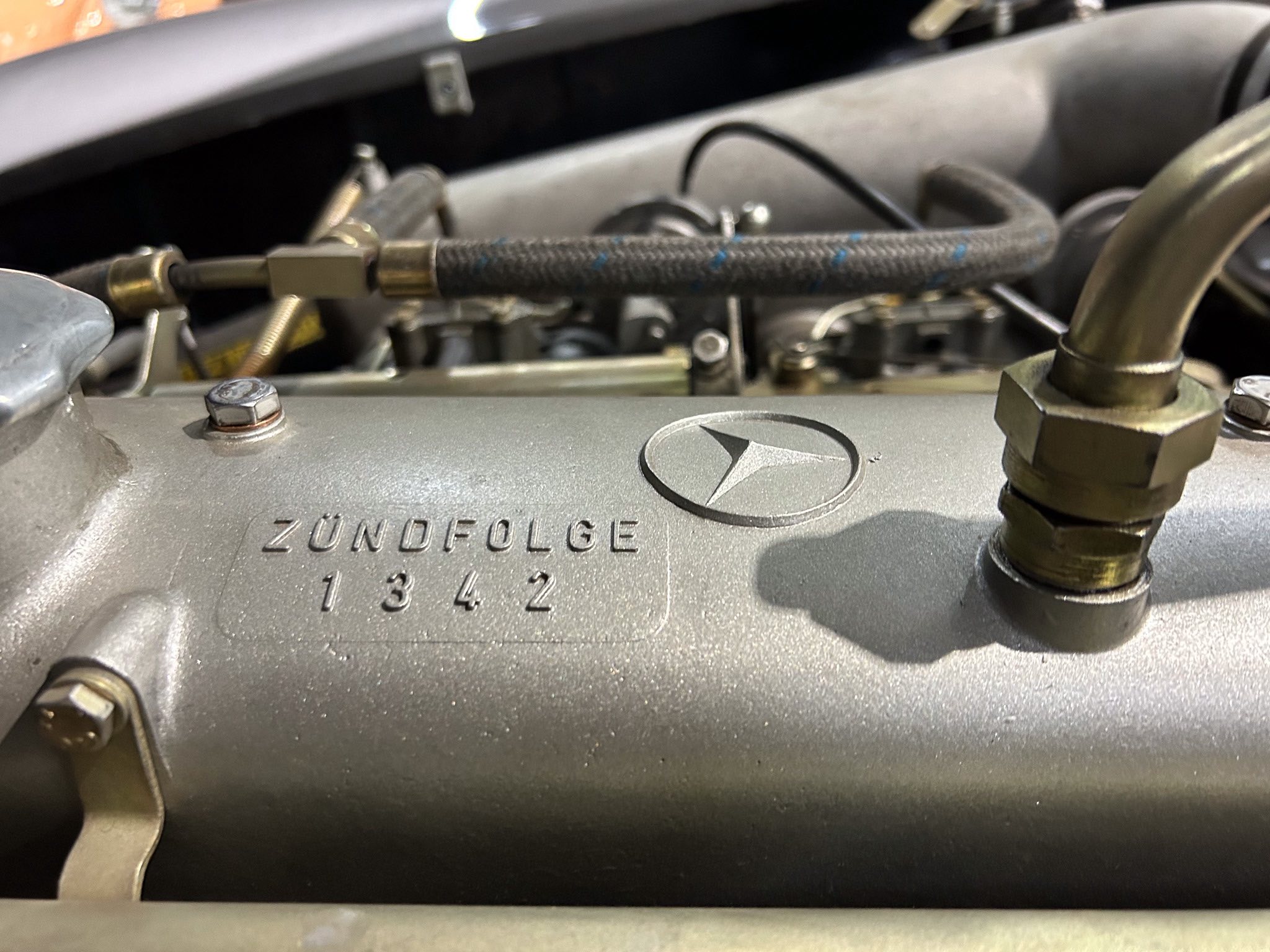 Close-up of a Mercedes engine cover showing ignition sequence markings 'ZUNDFOLGE 1 3 4 2' with nearby hoses and bolts.