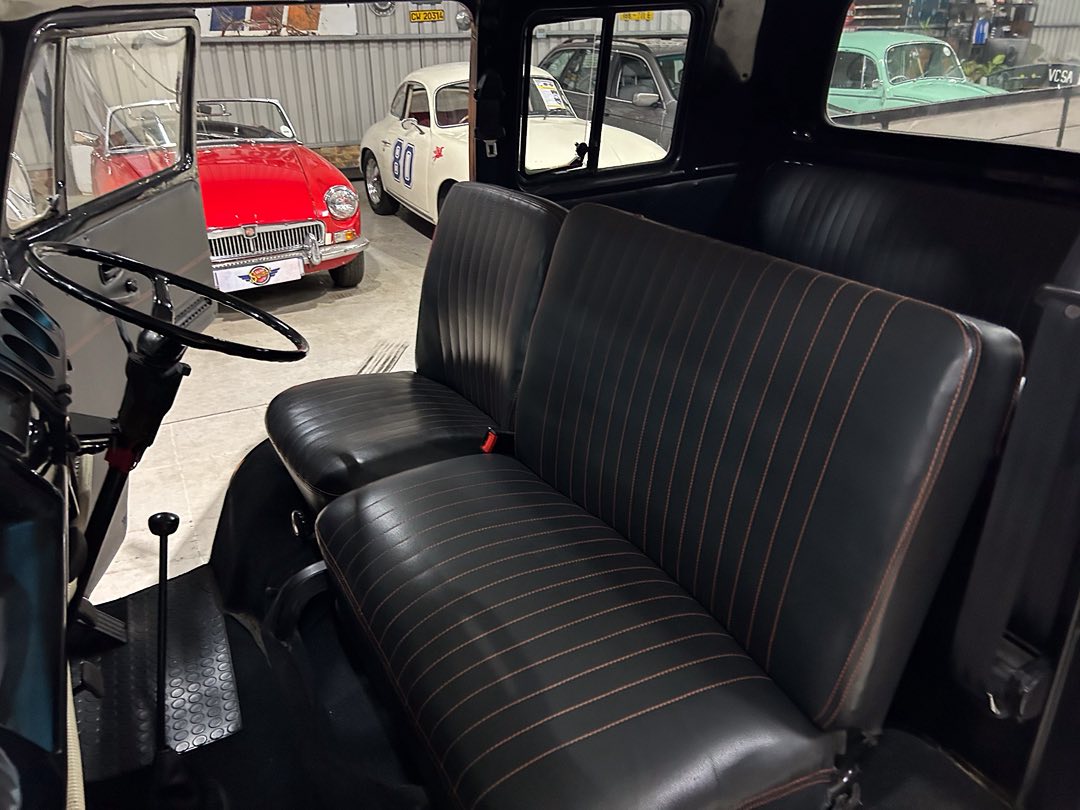 Interior of a vintage car with black leather seats and orange stitching, steering wheel, and gear lever; classic cars visible through the windows in a showroom.