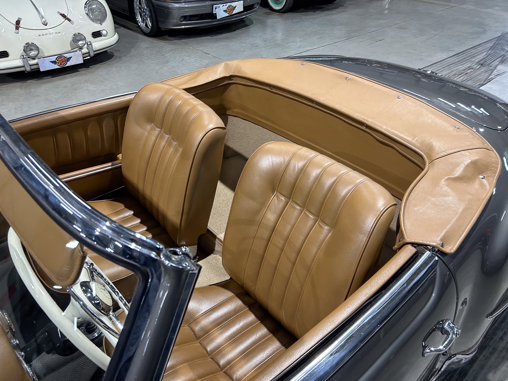 Tan leather interior of a vintage convertible with two front seats, open top, chrome trim, in a showroom setting with other classic cars in the background