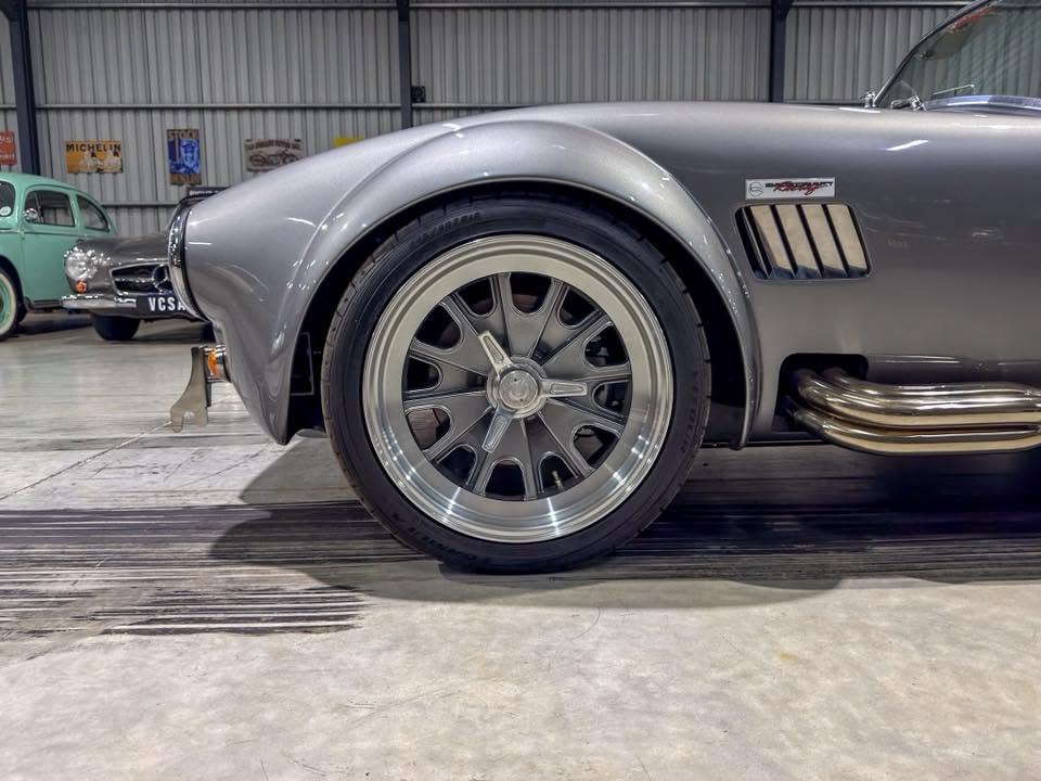 Close-up of front left wheel and fender of a silver vintage car with a multi-spoke alloy wheel in a showroom setting.
