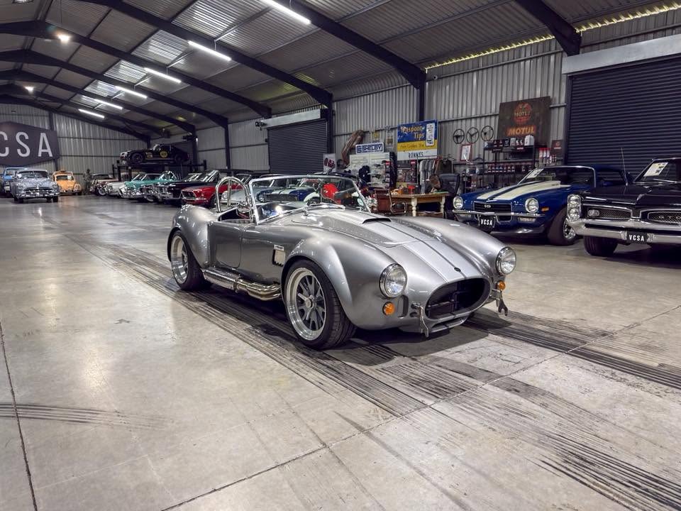 Silver Shelby Cobra–style roadster with exposed exhausts in a vintage car showroom in the foreground.