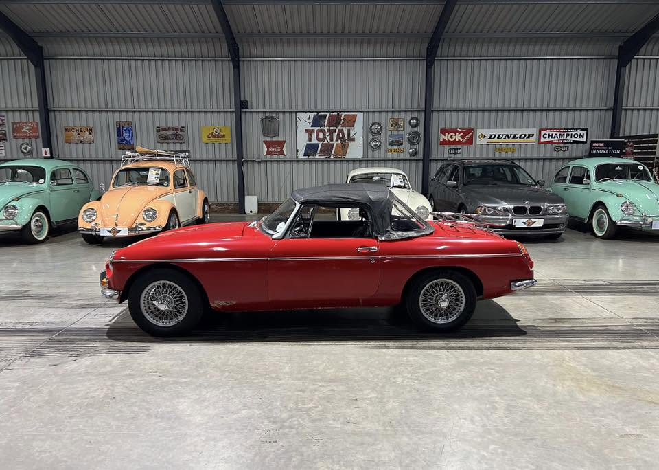 Red vintage convertible with a black soft top parked inside a classic car showroom, surrounded by other restored cars and retro banners on the wall.