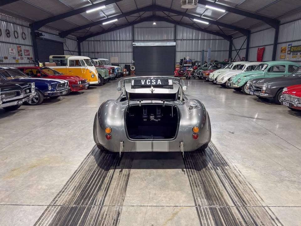 Rear view of a silver vintage race car with an open trunk in a classic car showroom, surrounded by colorful retro automobiles.