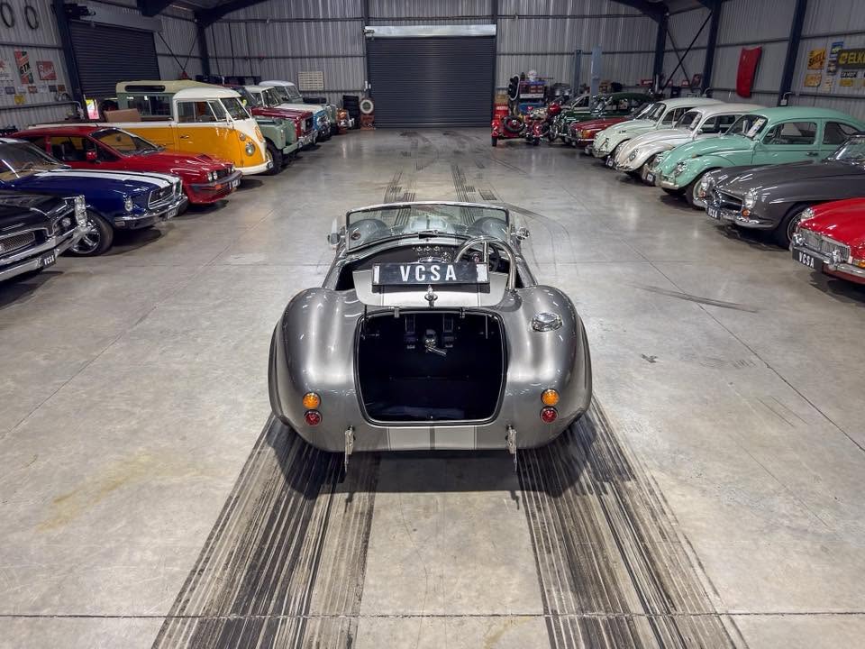 Rear view of a silver vintage open-top sports car in a classic car showroom, flanked by rows of colorful vintage cars on both sides.
