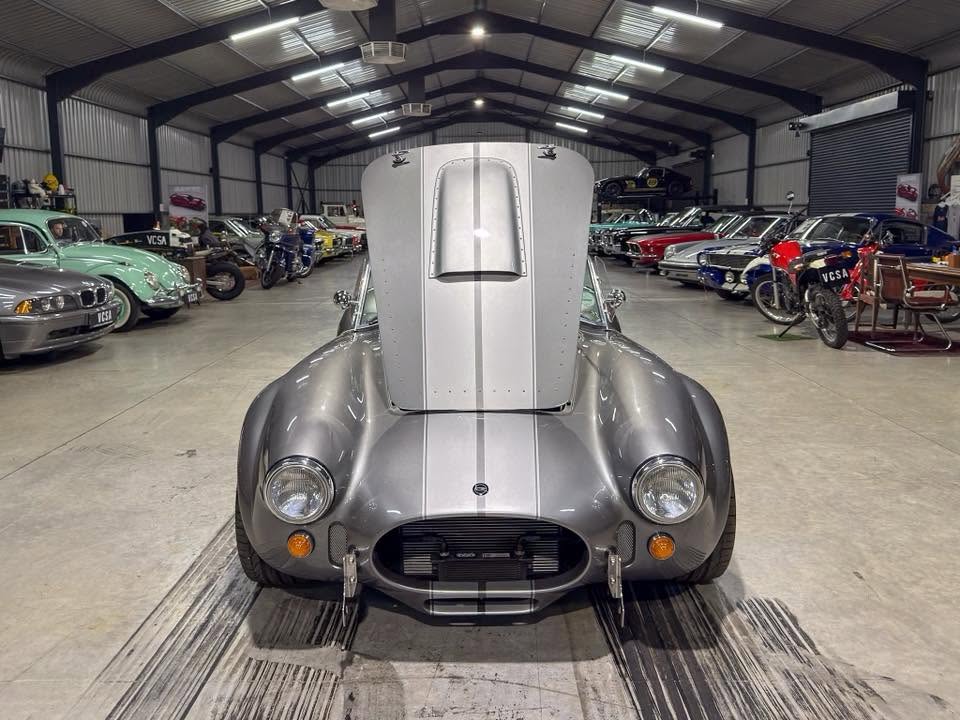 Front view of a silver vintage race car with a tall raised hood in a large classic car showroom.