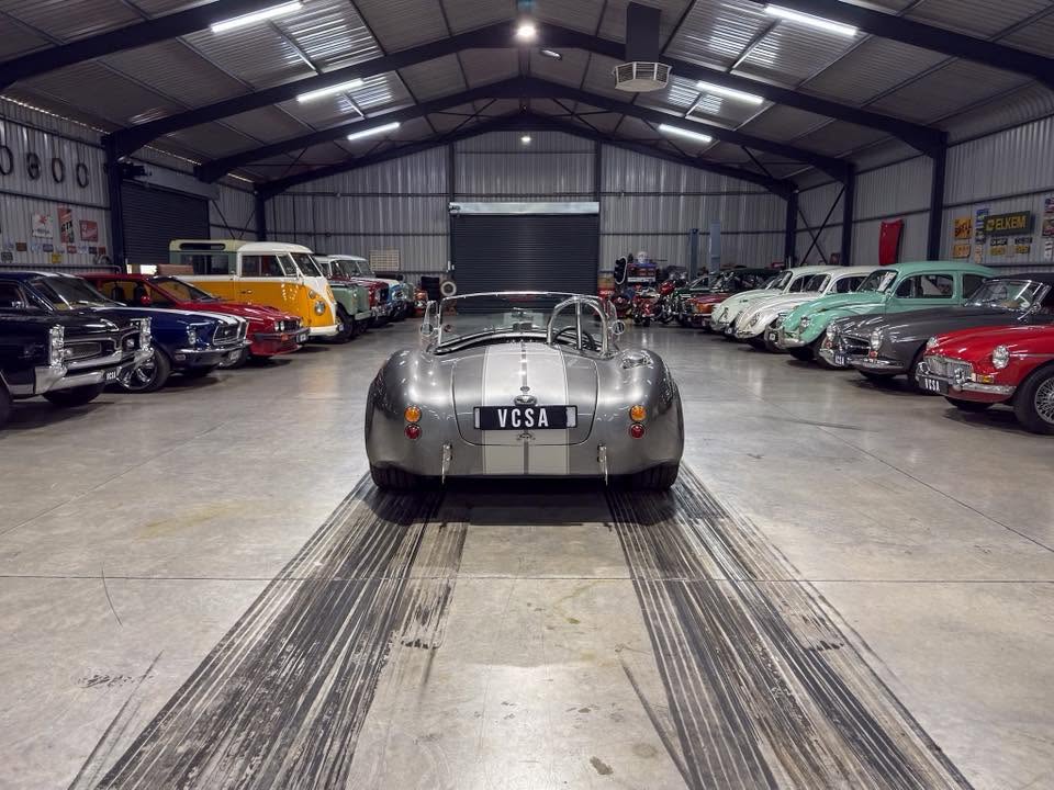 Silver vintage roadster parked center in a spacious indoor car showroom, surrounded by classic cars on both sides.