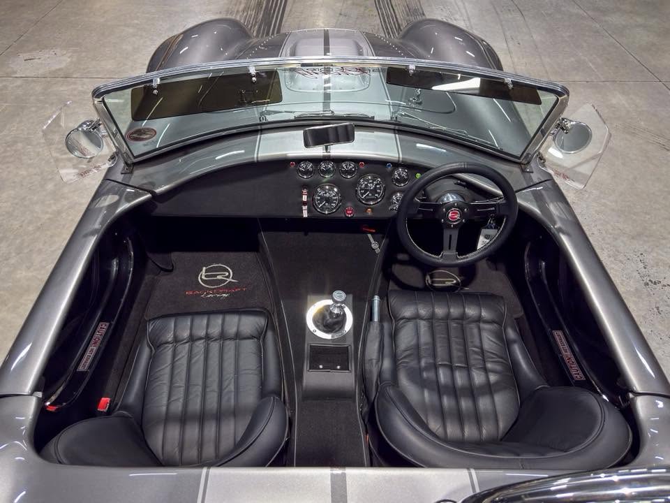 Top-down view of a silver vintage sports car interior with black leather seats, dashboard gauges, and a three-spoke steering wheel in front of the driver and passenger.