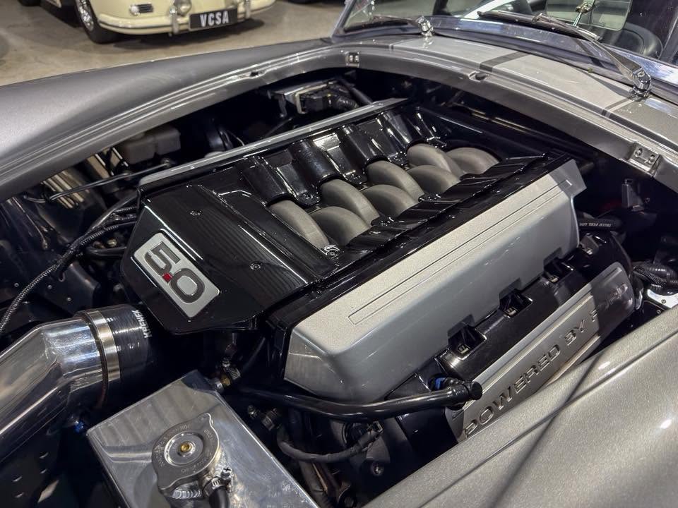 Under-hood view of a high-performance car engine in a showroom, featuring a prominent silver 6.0 badge on a black intake manifold and polished silver components behind the hood sill. It’s a V8 powerplant with multiple intake runners visible.