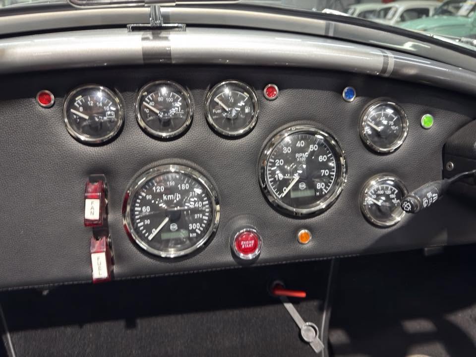 Vintage car dashboard with multiple round gauges, including speedometer in km/h and tachometer, red start button, and toggle switches on black vinyl panel