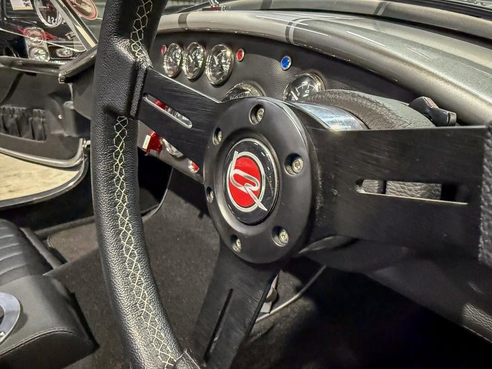 Inside a classic car: black three-spoke steering wheel with a red center emblem, chrome bolts, and a cluster of round gauges on the dashboard.