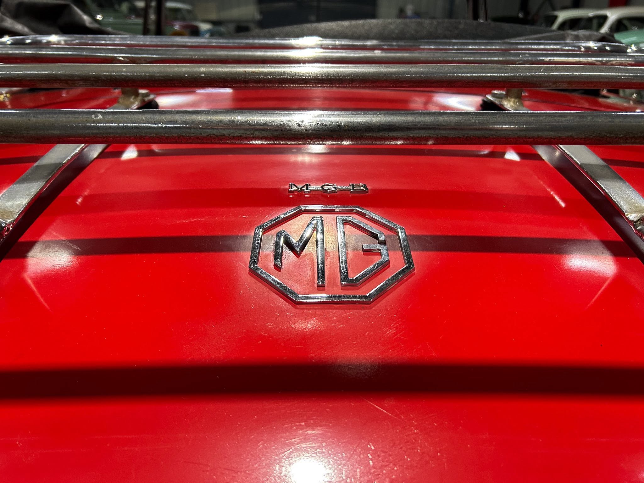 Close-up of a chrome MG emblem on a glossy red car surface with horizontal chrome bars across the image