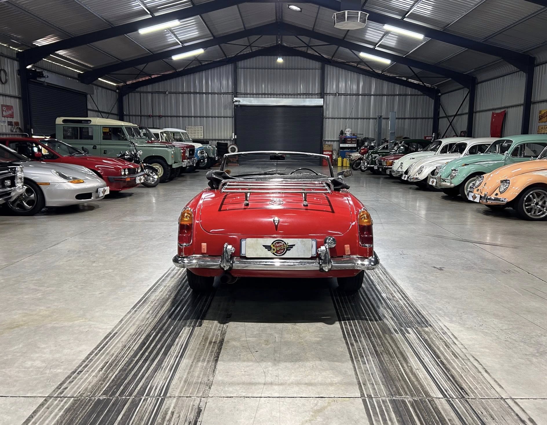 Rear view of a red vintage convertible centered in a spacious showroom, flanked by rows of classic cars on both sides inside a metal industrial building.