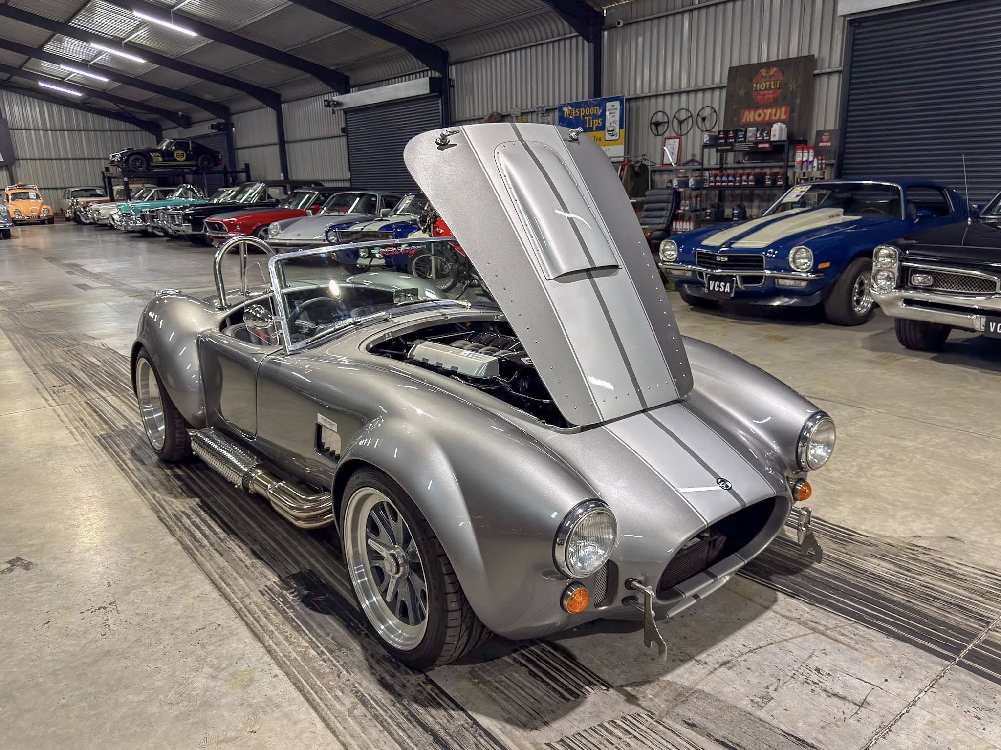 Classic car showroom with a silver vintage roadster in the foreground, hood raised for inspection and other vintage cars lined up in the background under a metal-roofed warehouse.