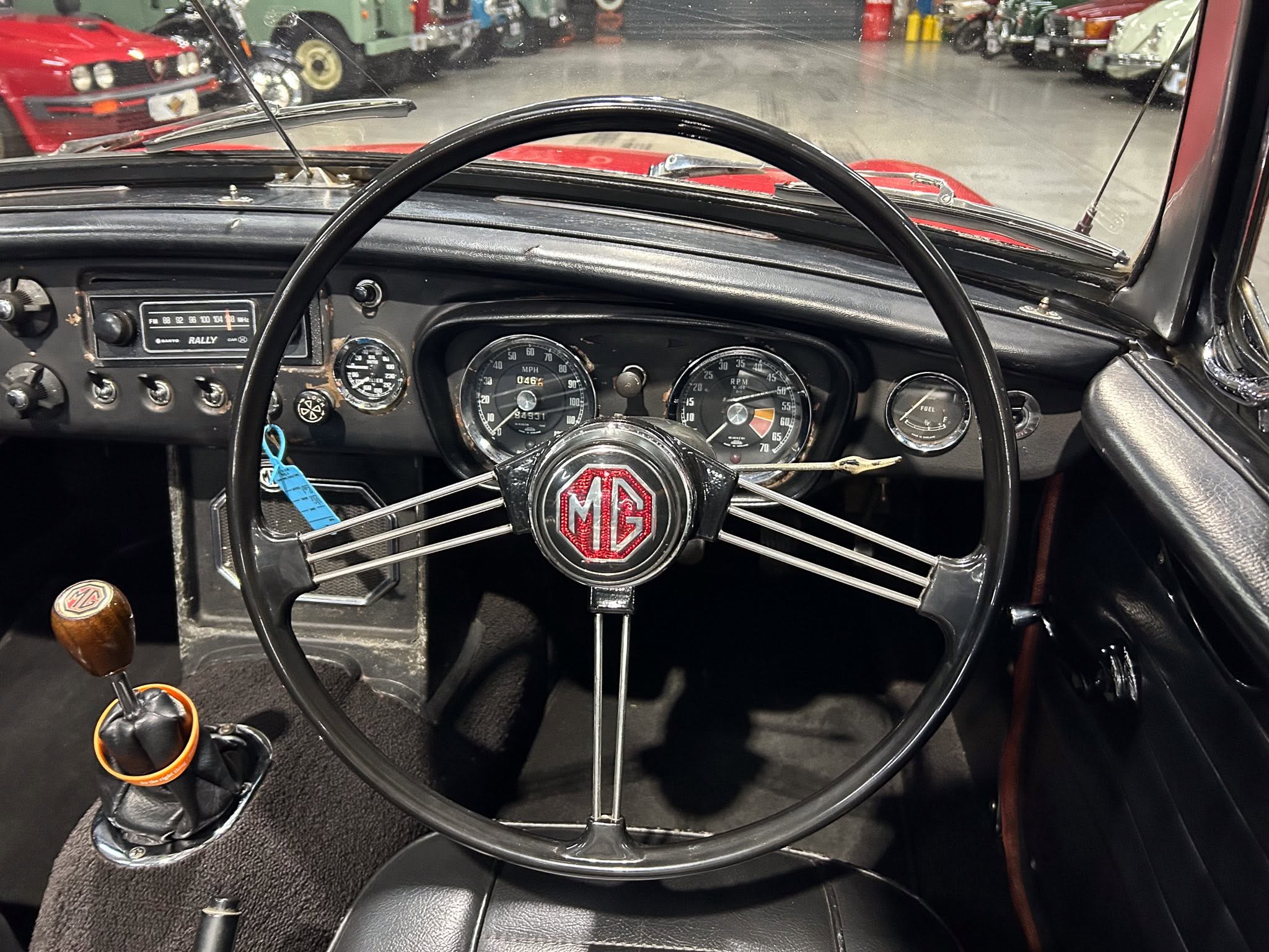 Interior view of a vintage MG car showing a large spoke steering wheel, analog gauges, and a gear lever with a blue tag nearby inside a showroom.