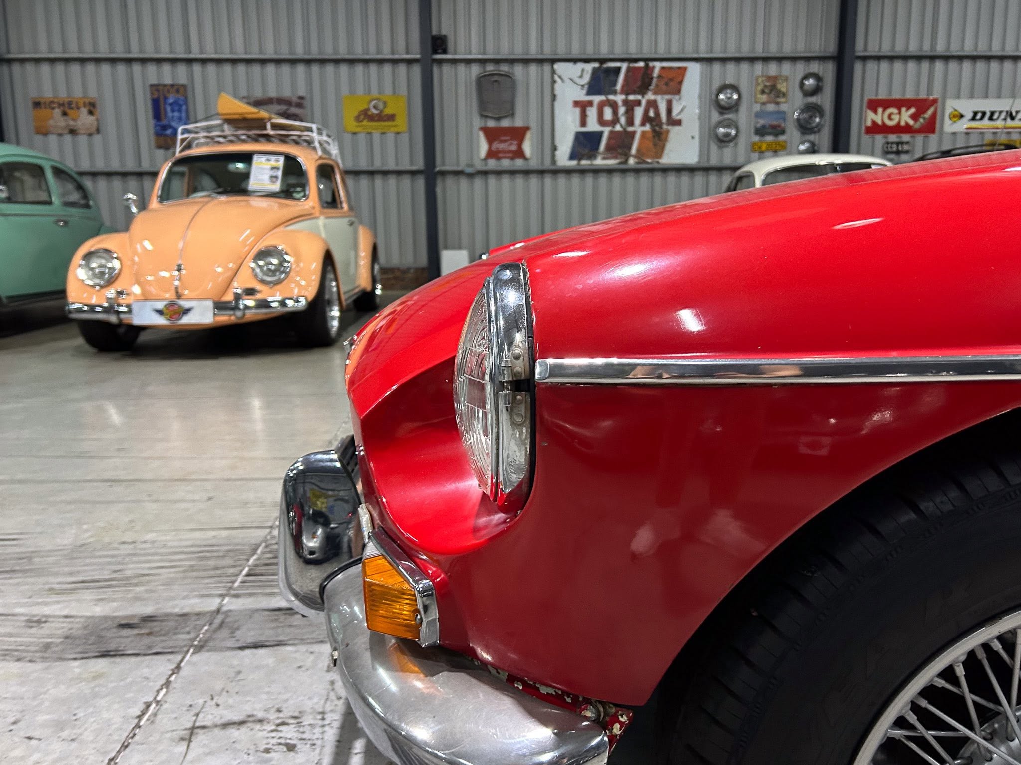 Close-up of a shiny red vintage car's front fender and headlight, with a pale peach classic car in the background inside a showroom.