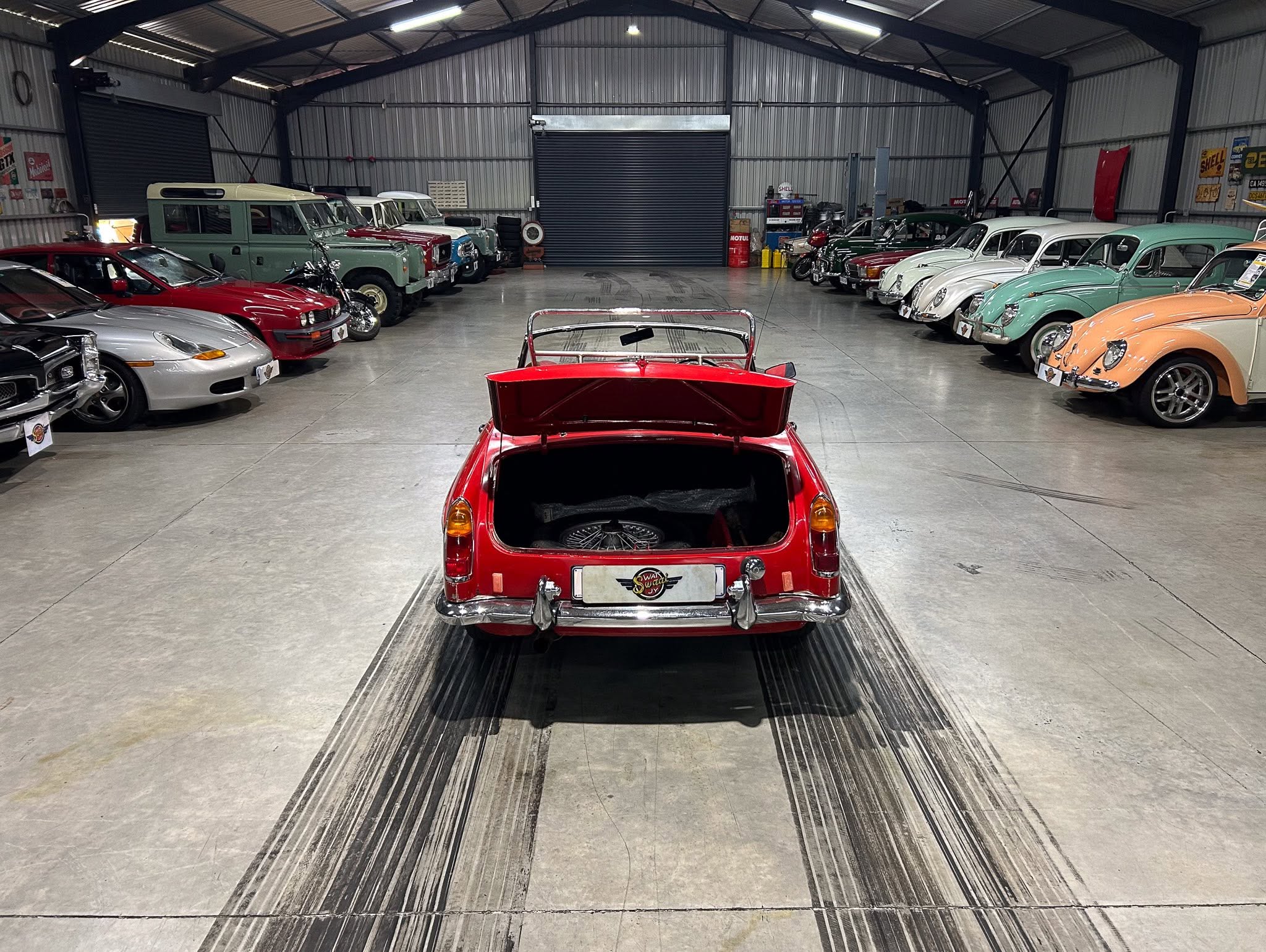Red vintage car with open trunk parked center, flanked by rows of classic cars in a large showroom hall, metal walls in view.