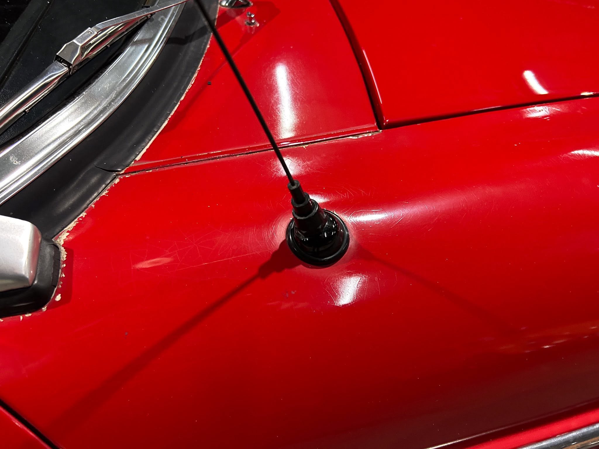 Close-up of a shiny red car body showing chipped paint along the door seam with a black antenna in the foreground.