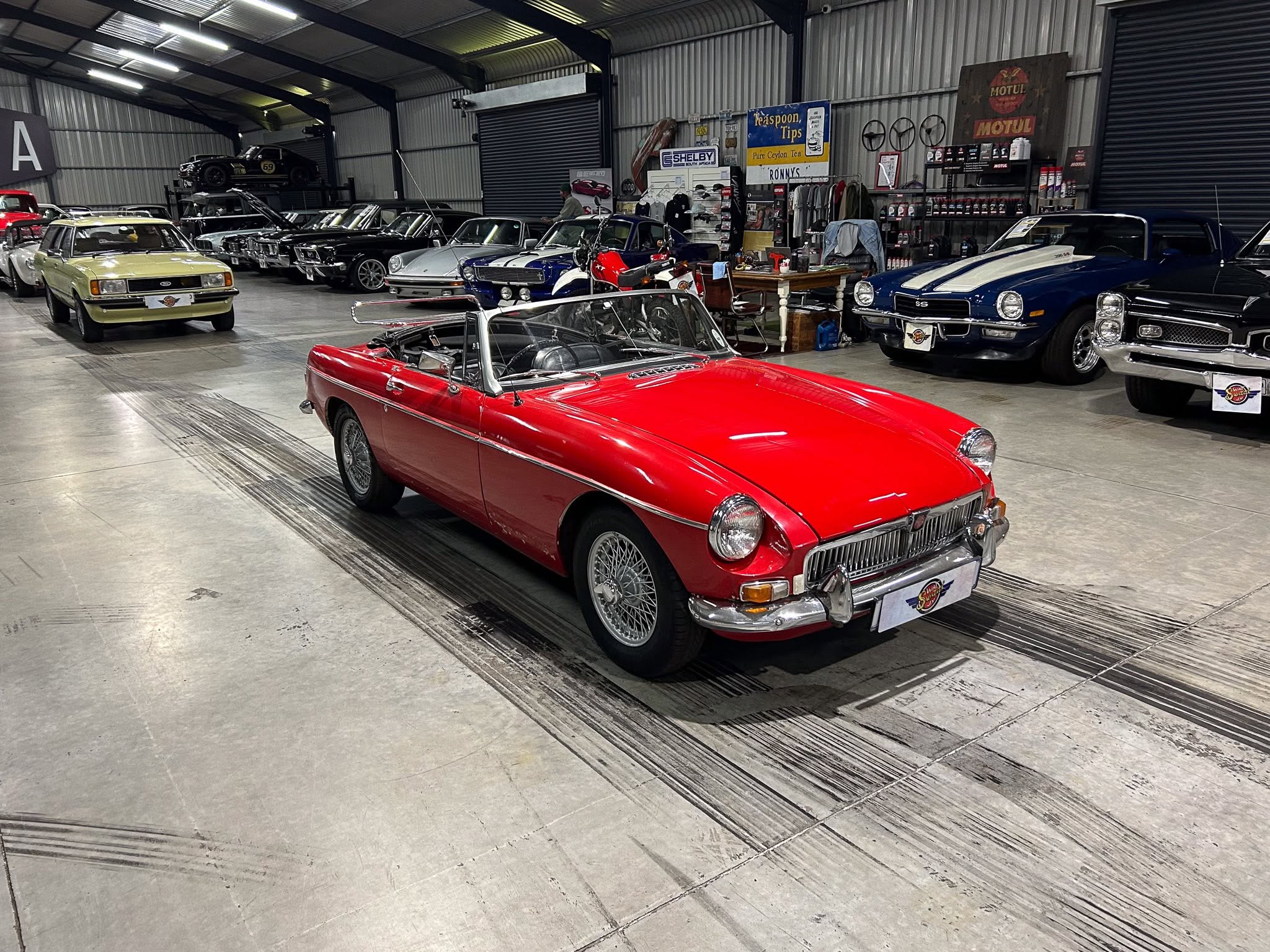 Inside a large garage filled with classic cars lined up along the wall, including a red convertible in the foreground and a yellow sedan to the left