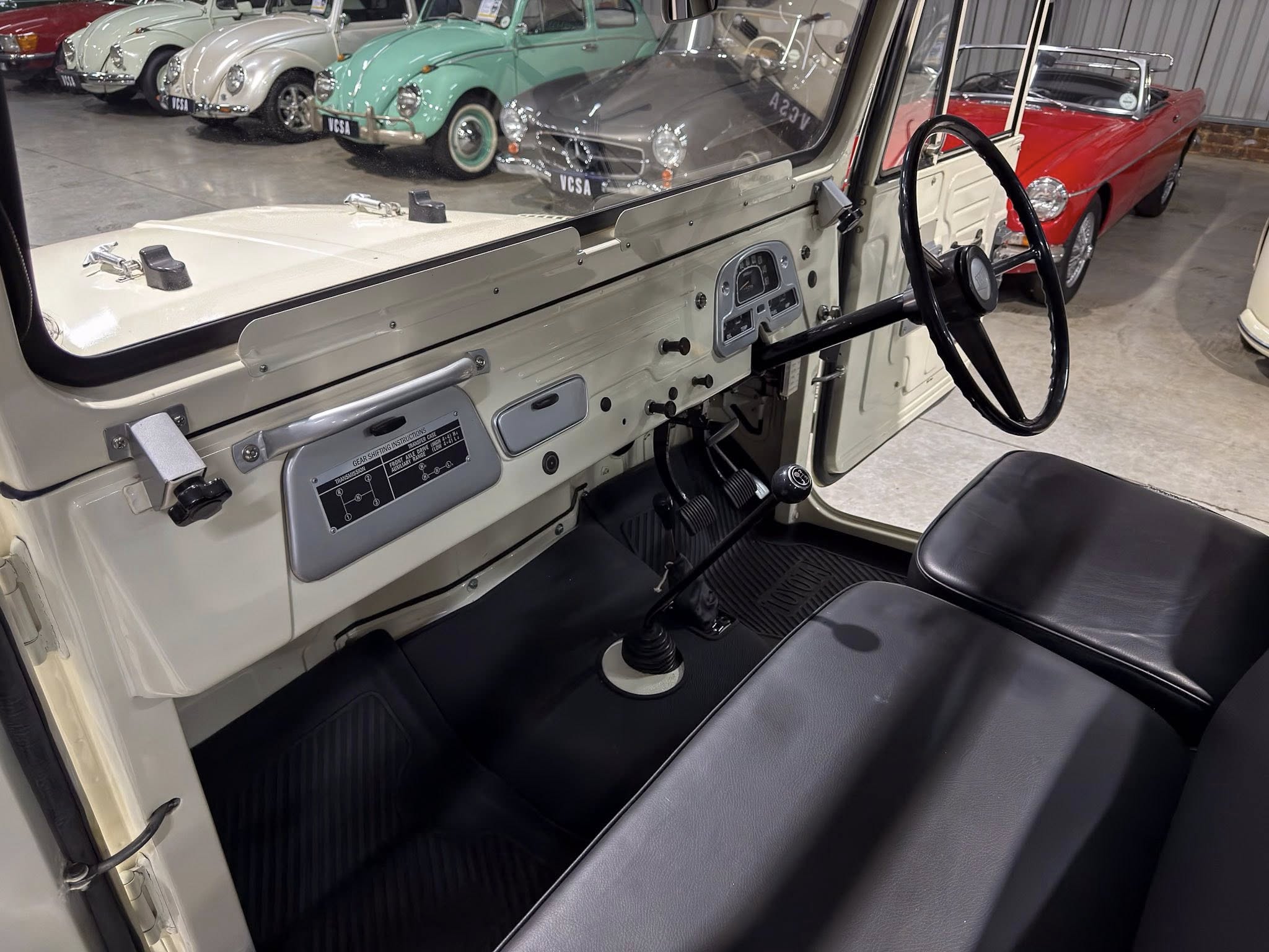 Interior view of a vintage right-hand-drive SUV, showing a cream-colored dashboard, large black steering wheel, and gear levers with a row of classic cars in a showroom behind.