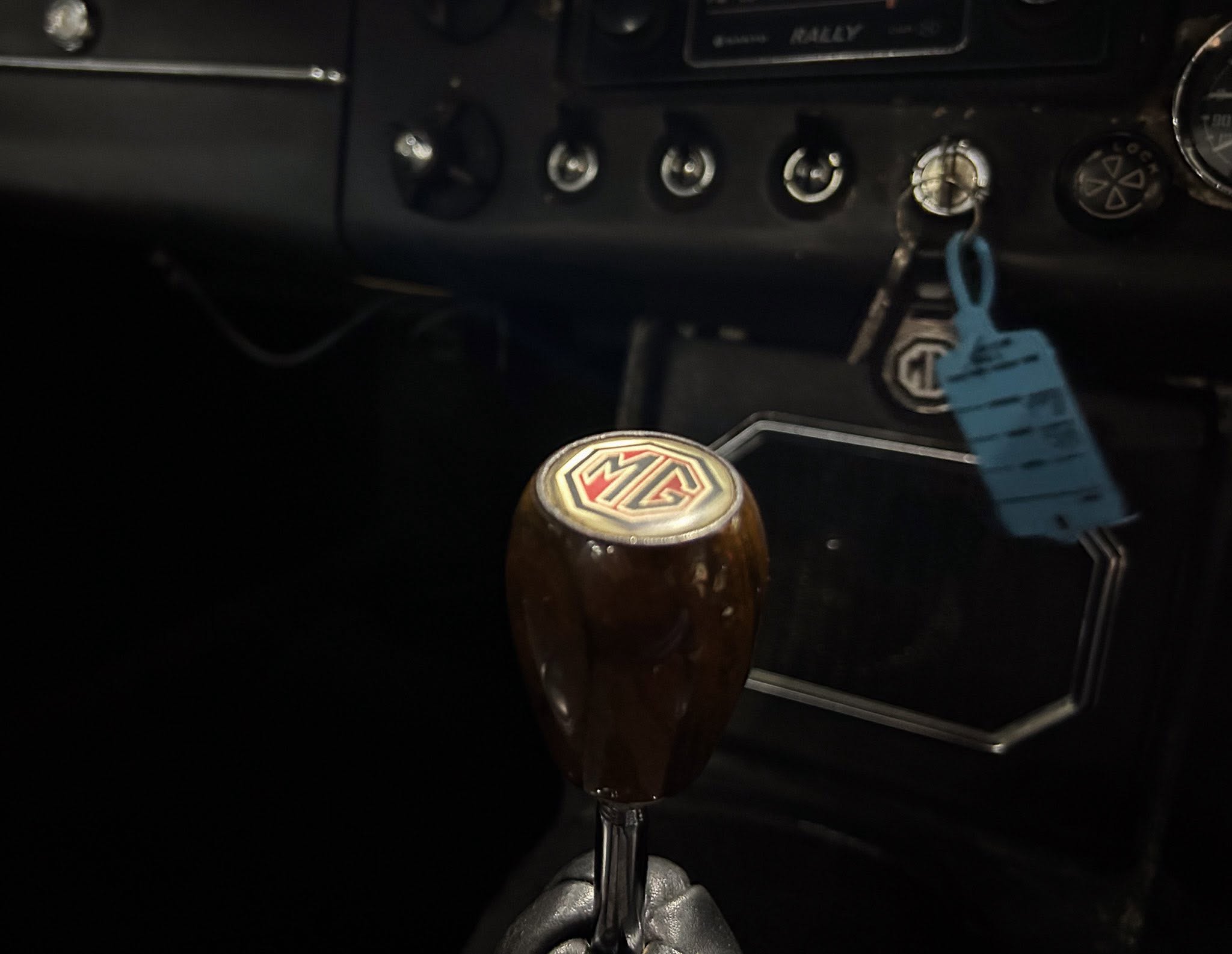 Close-up of a vintage car gear lever with an MG emblem on the knob, ignition keys in the dash, and a dark dashboard.