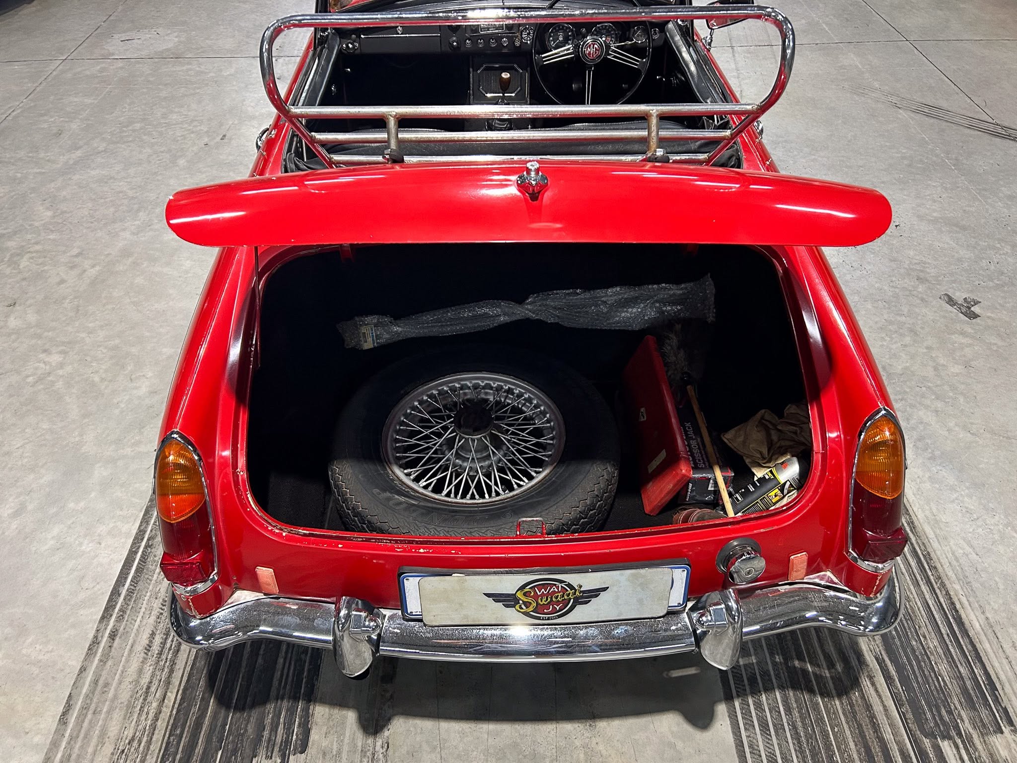 Rear view of a red vintage MG with the trunk open, spare wheel visible inside and chrome bumper visible below common tail lights on each side.