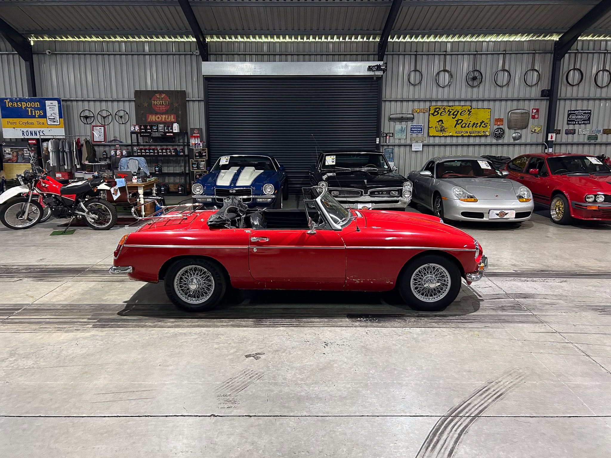 Indoor showroom or garage with a red vintage convertible in the foreground and several classic cars parked along the back wall, metal walls, and wall-mounted bike wheels/signs visible in the background