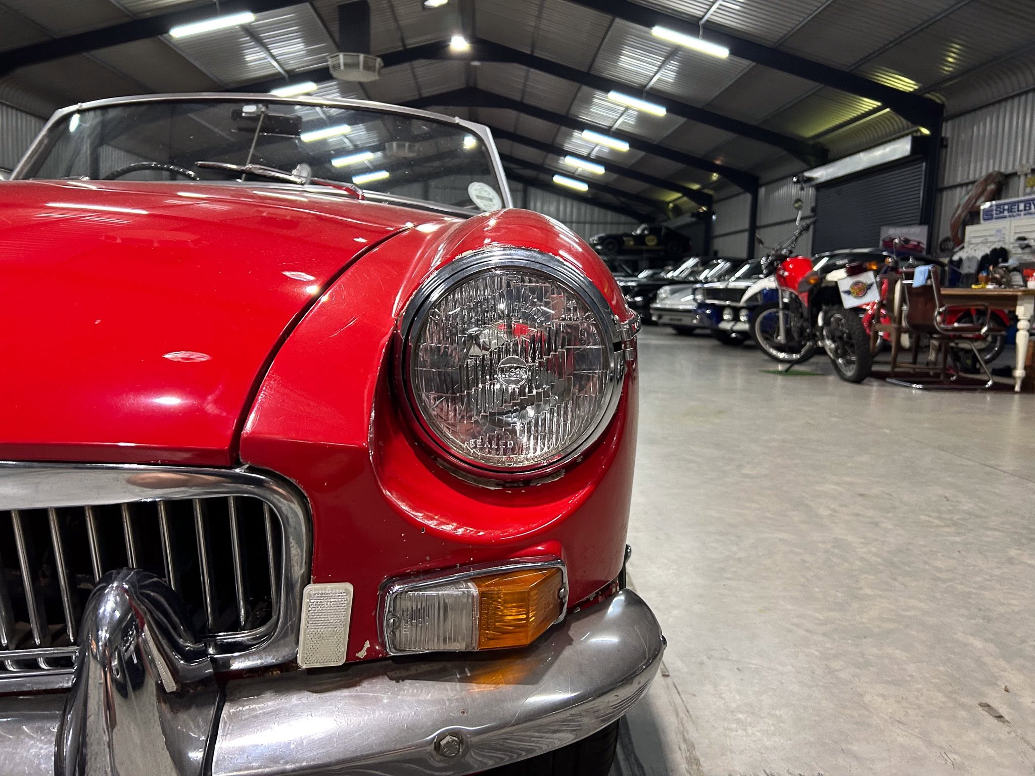 Close-up of the red front end and round headlight of a vintage car inside a showroom, with other vehicles in the background and showroom lights overhead.