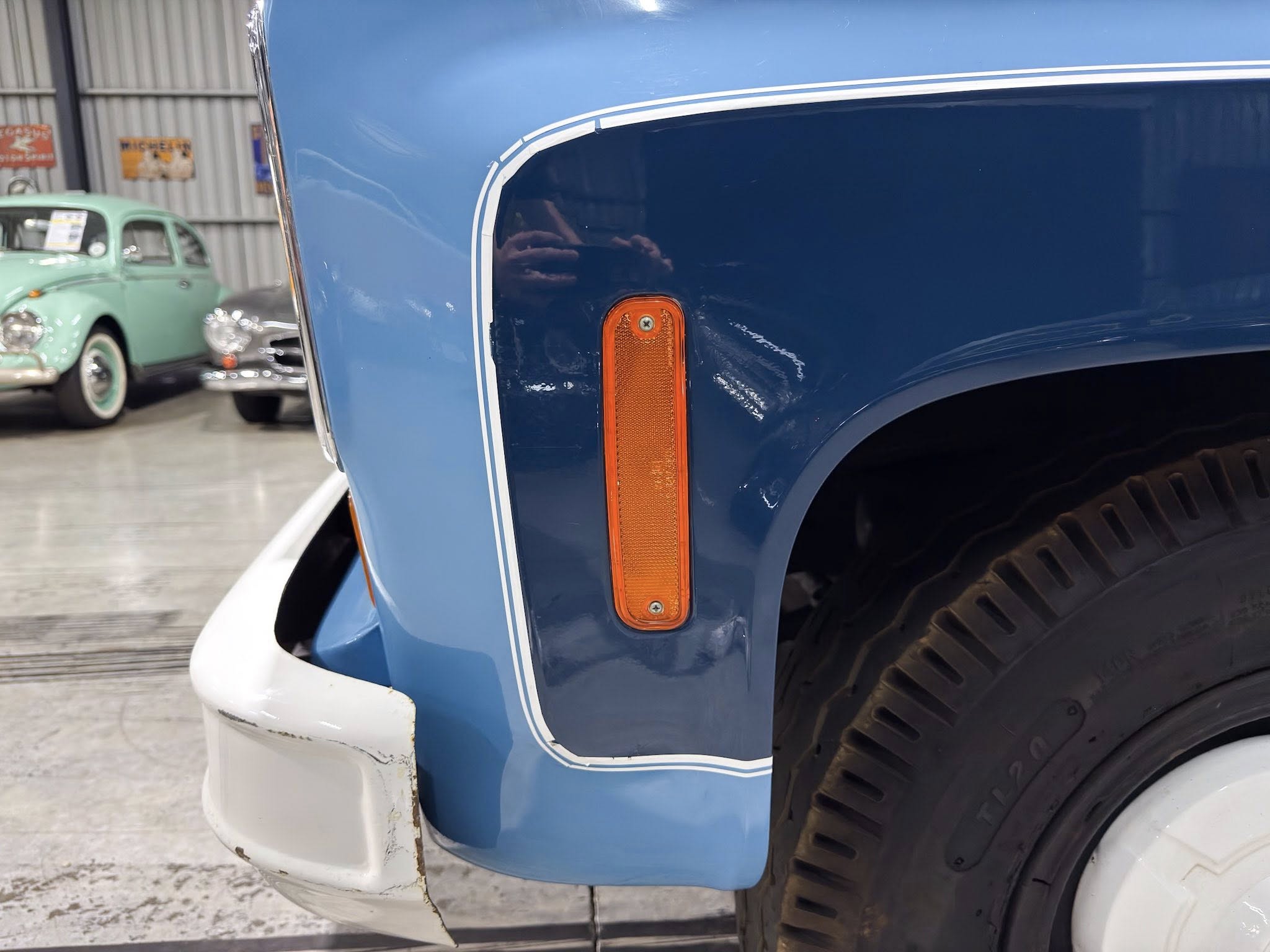 Close-up of a blue vintage van's front fender with an orange reflector, white bumper, and a tire visible; background shows other classic cars in a workshop.