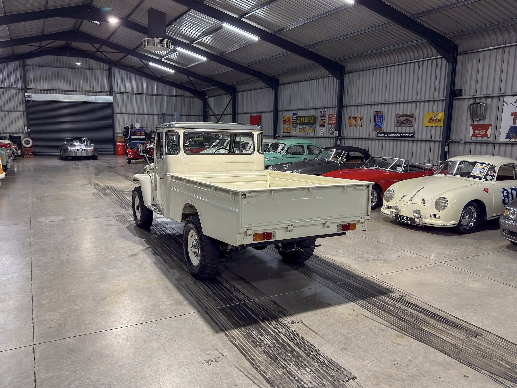 Indoor showroom of vintage cars: white pickup in foreground with a row of classic vehicles along the wall under metal ceiling panels and retro posters.