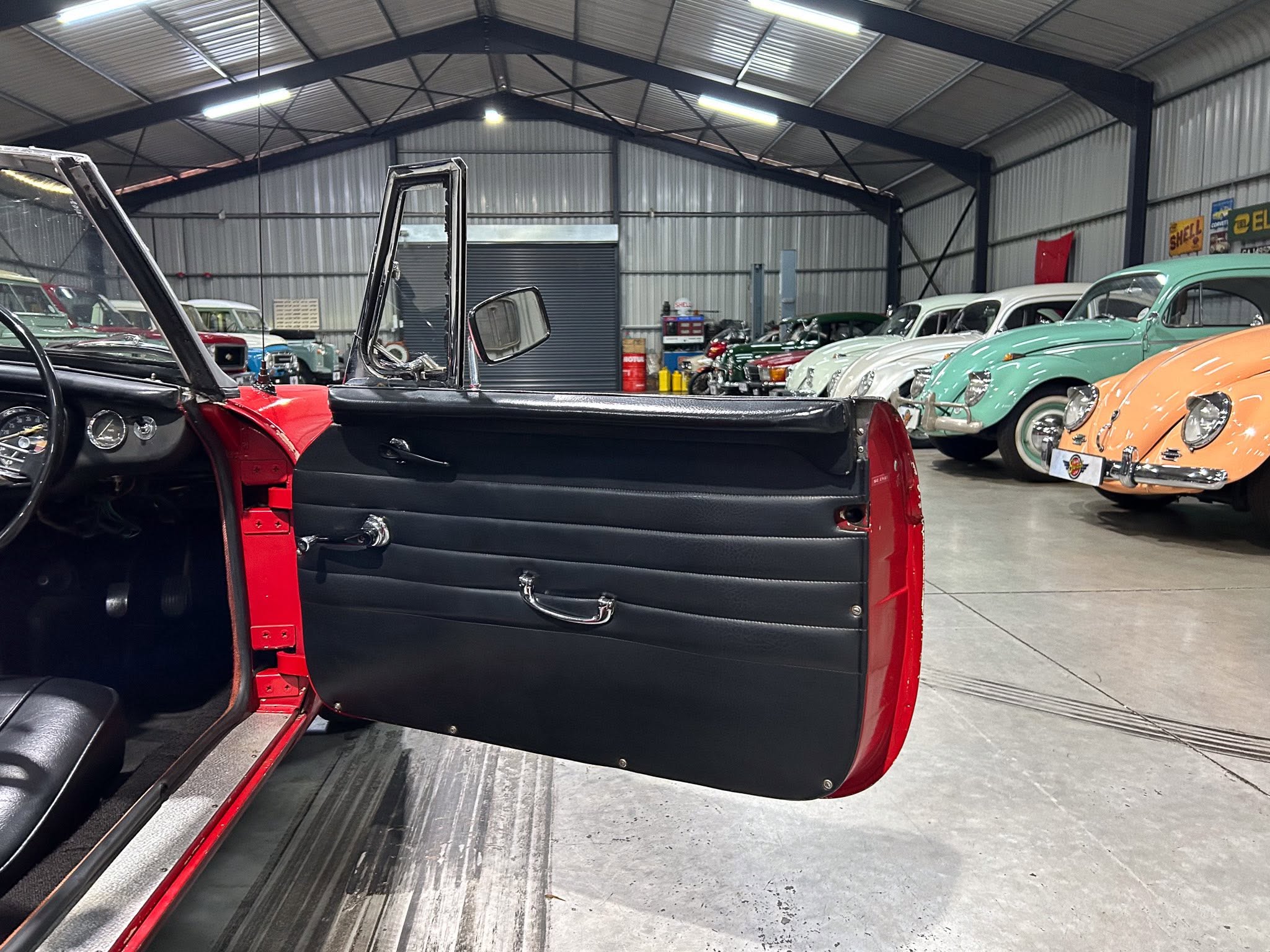 Open red classic car door in foreground with black interior; showroom filled with pastel-colored vintage cars under industrial lighting in back.