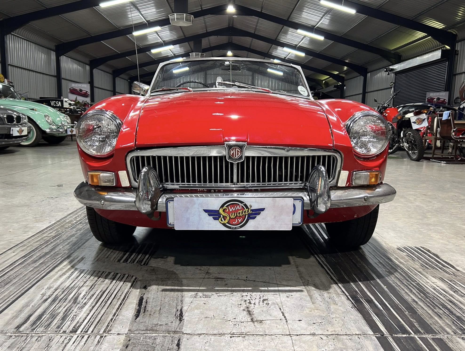 Front view of a red MG classic car on display in a showroom, with other vintage cars visible in the background.