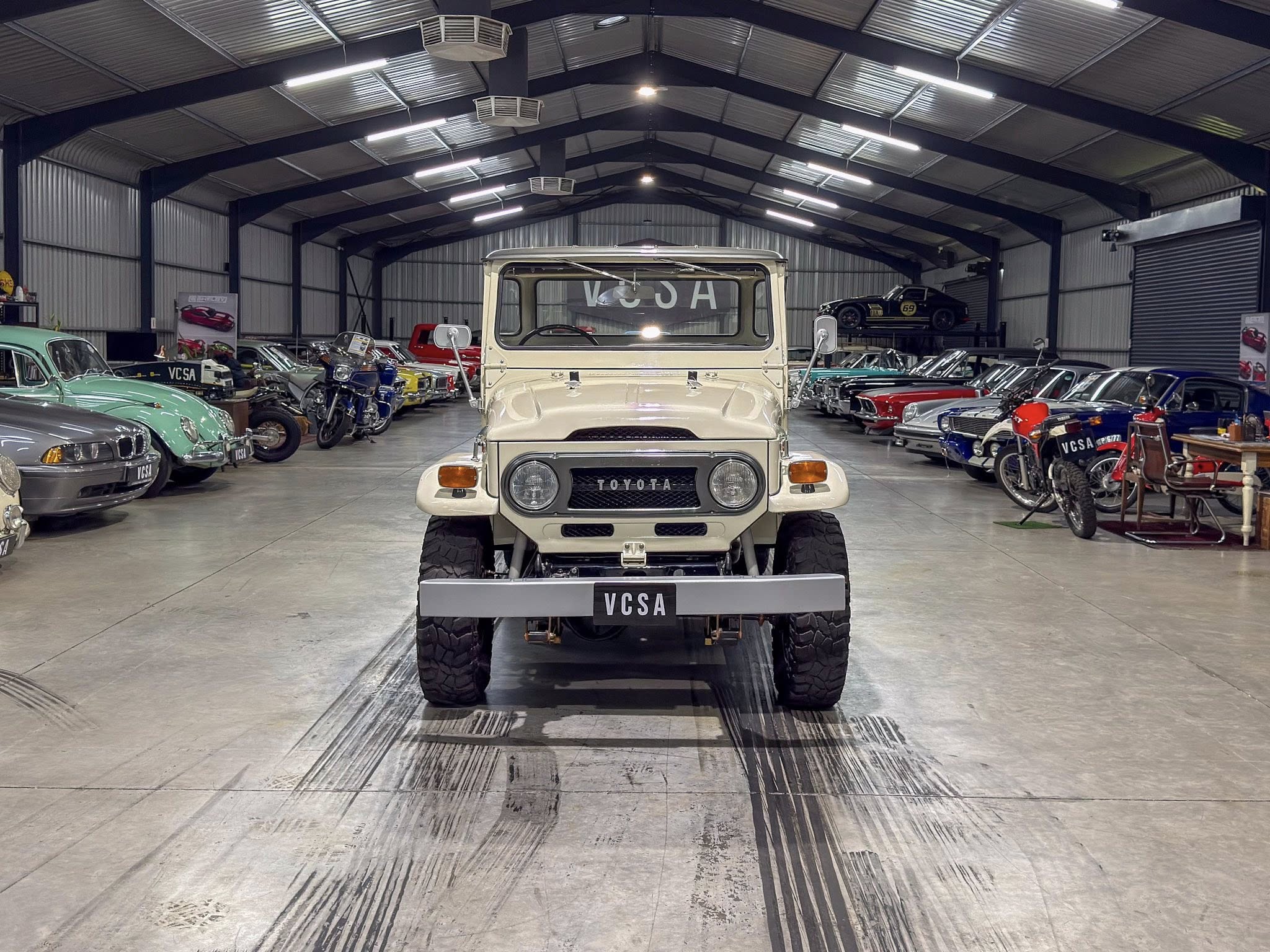 Indoor showroom of a vintage car collection; a beige Toyota Land Cruiser sits centered, flanked by classic cars and motorcycles under bright warehouse lights.