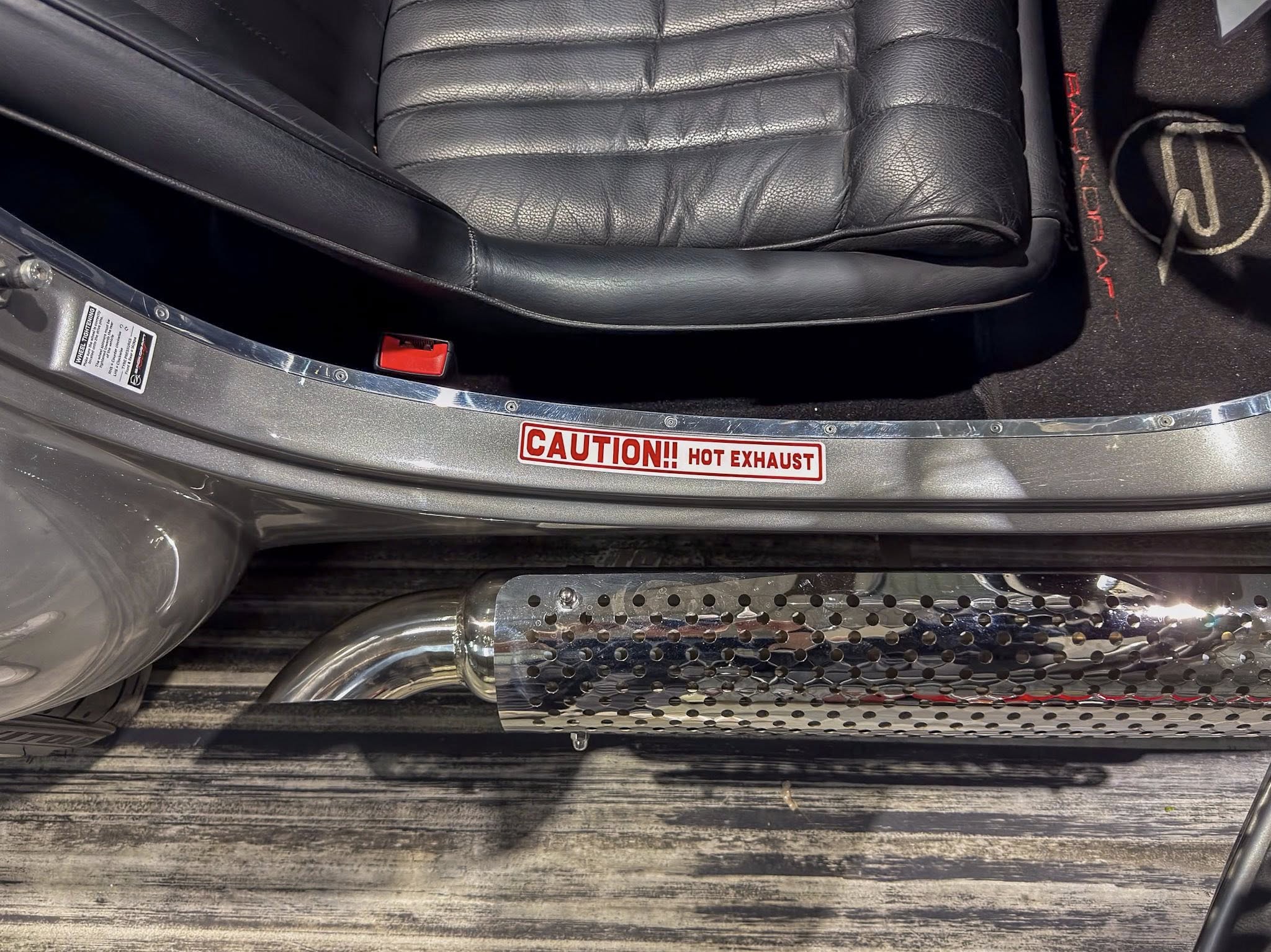 Close-up of a black leather car seat and red seat belt buckle at the door sill, with a metal heat shield and a 'CAUTION! HOT EXHAUST' label visible.