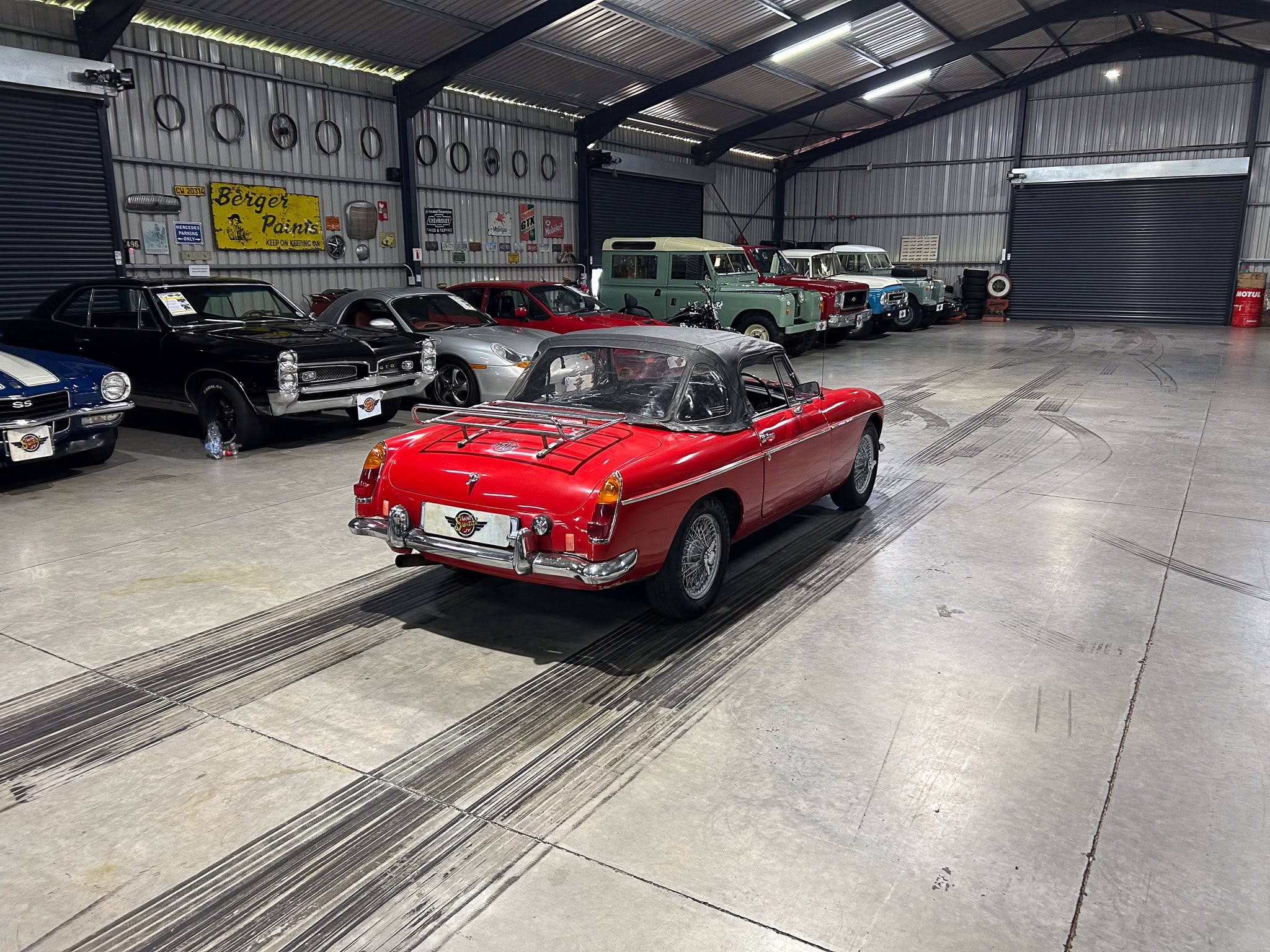 Inside a large industrial warehouse showroom with a row of classic cars, including a red convertible in the foreground and vintage vans in the back.