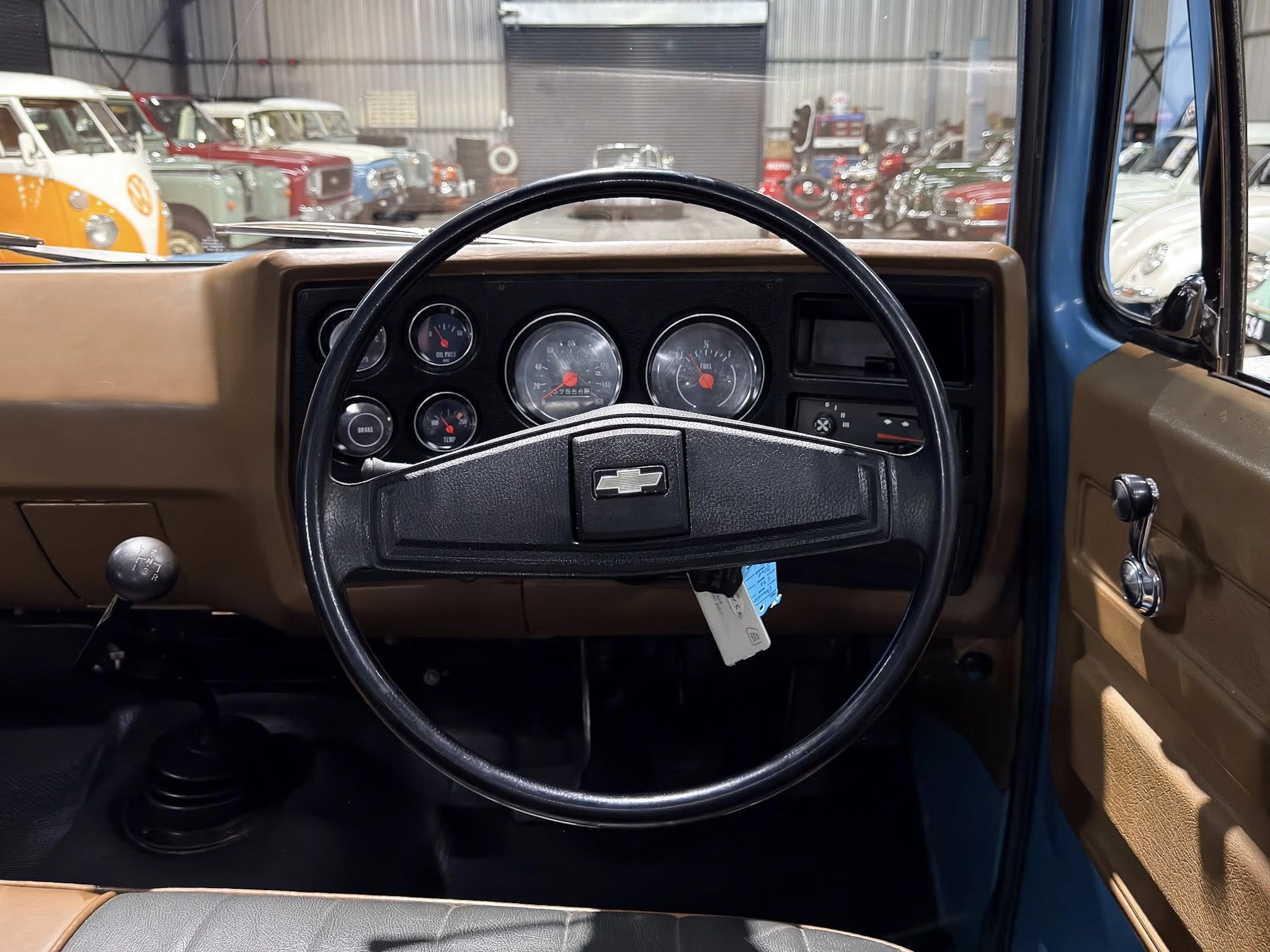 Driver’s-eye view of a vintage car dashboard with a black steering wheel and tan interior, and a row of classic cars in a workshop background.