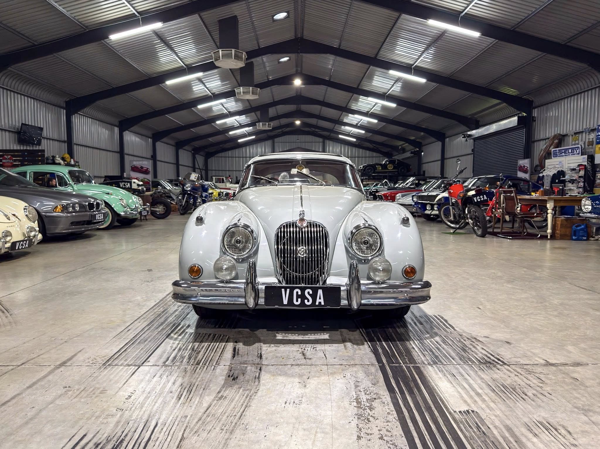Front-and-center silver vintage car inside a large showroom with other classic cars along the walls and ceiling lighting visible behind.