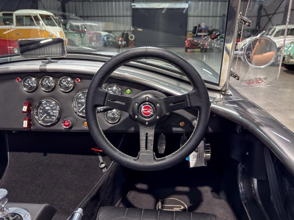 Interior of a vintage car: black three-spoke steering wheel, analog gauges, and metal dashboard in a showroom while viewed from the driver’s seat.