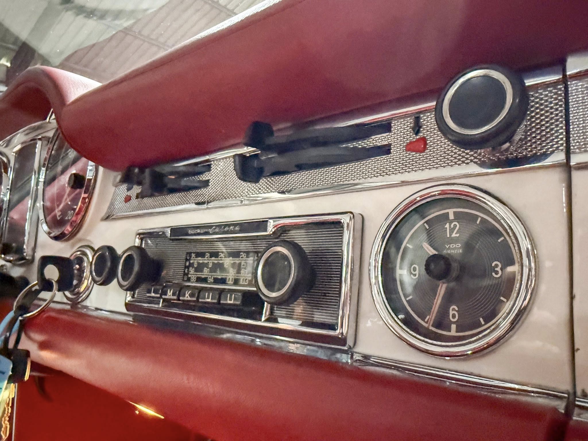 Close-up of a vintage car dashboard showing a large round tachometer and chrome trim with a radio and red padded interior behind it in a classic car cabin