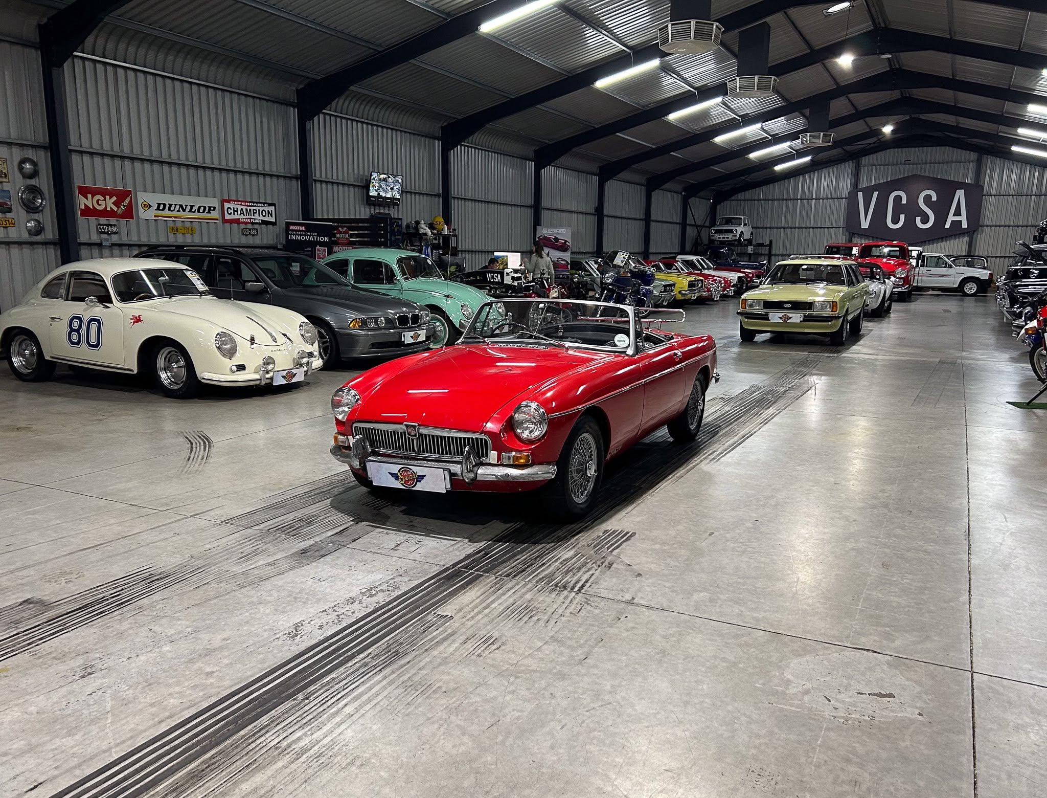 Indoor car showroom with a row of vintage cars on display; bright red convertible at the forefront and a cream numbered '80' car on the left, VCSA sign on the right wall.