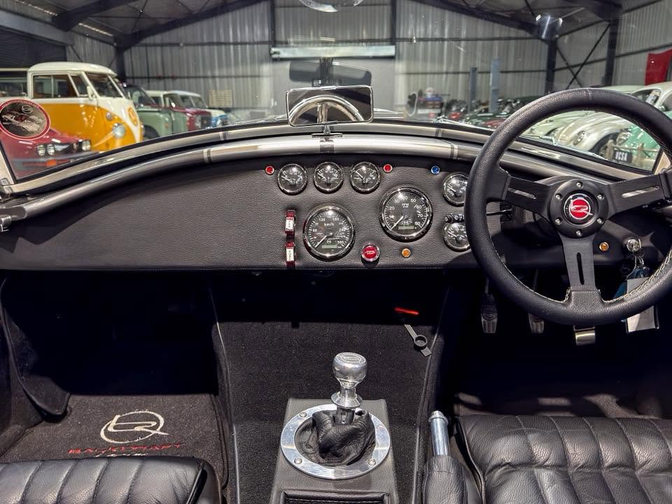 Interior of a classic car showing a vintage gauge cluster, black dash, and three-spoke steering wheel in a showroom with other vintage cars outside.