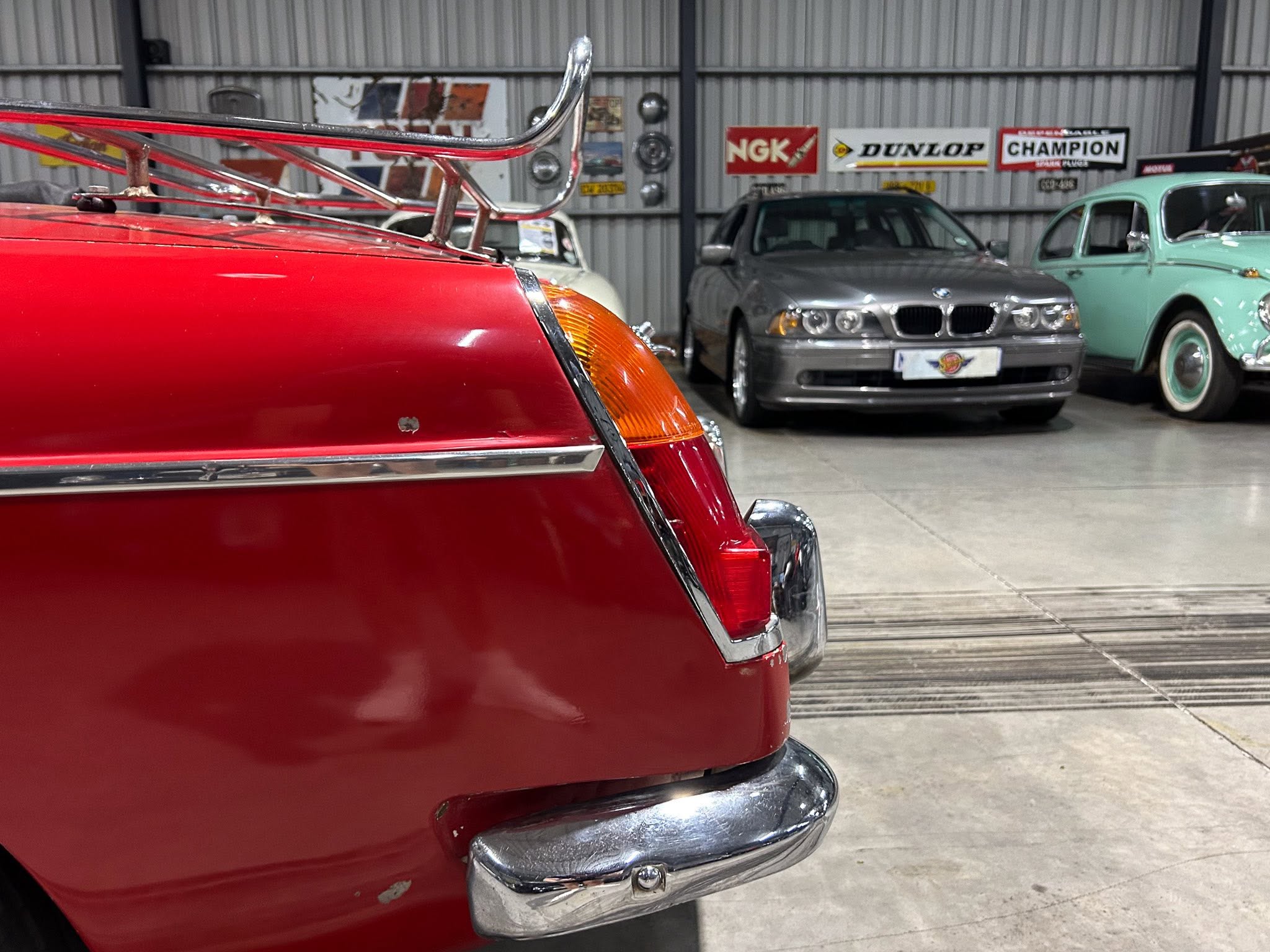 Close-up of a red car's front end with chrome bumper, orange signal light, indoors showroom with other classic cars in the background.