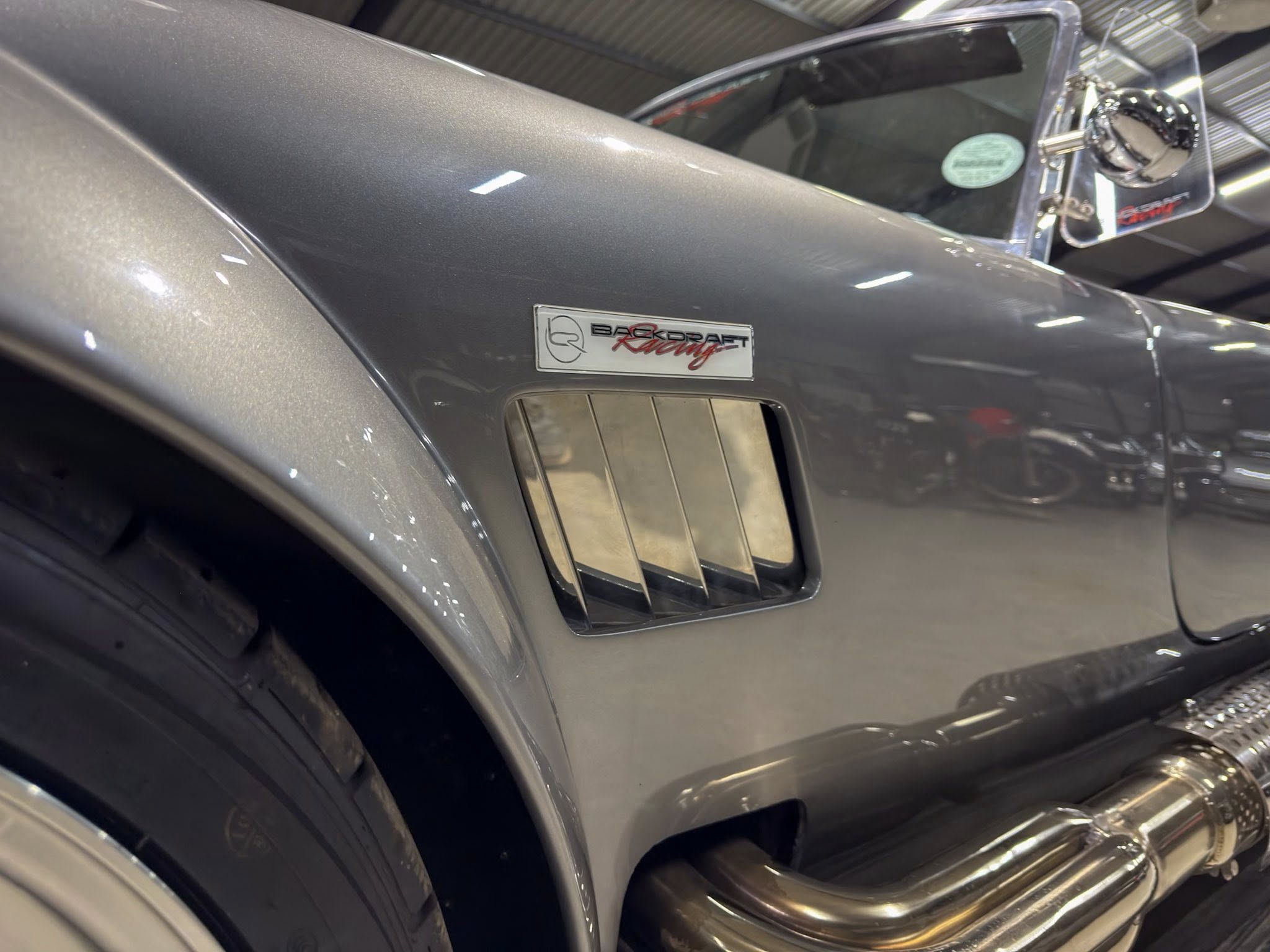 Close-up of a gray racing car's side, showing a Backdraft Racing badge on the door panel and chrome trim around a vent window.