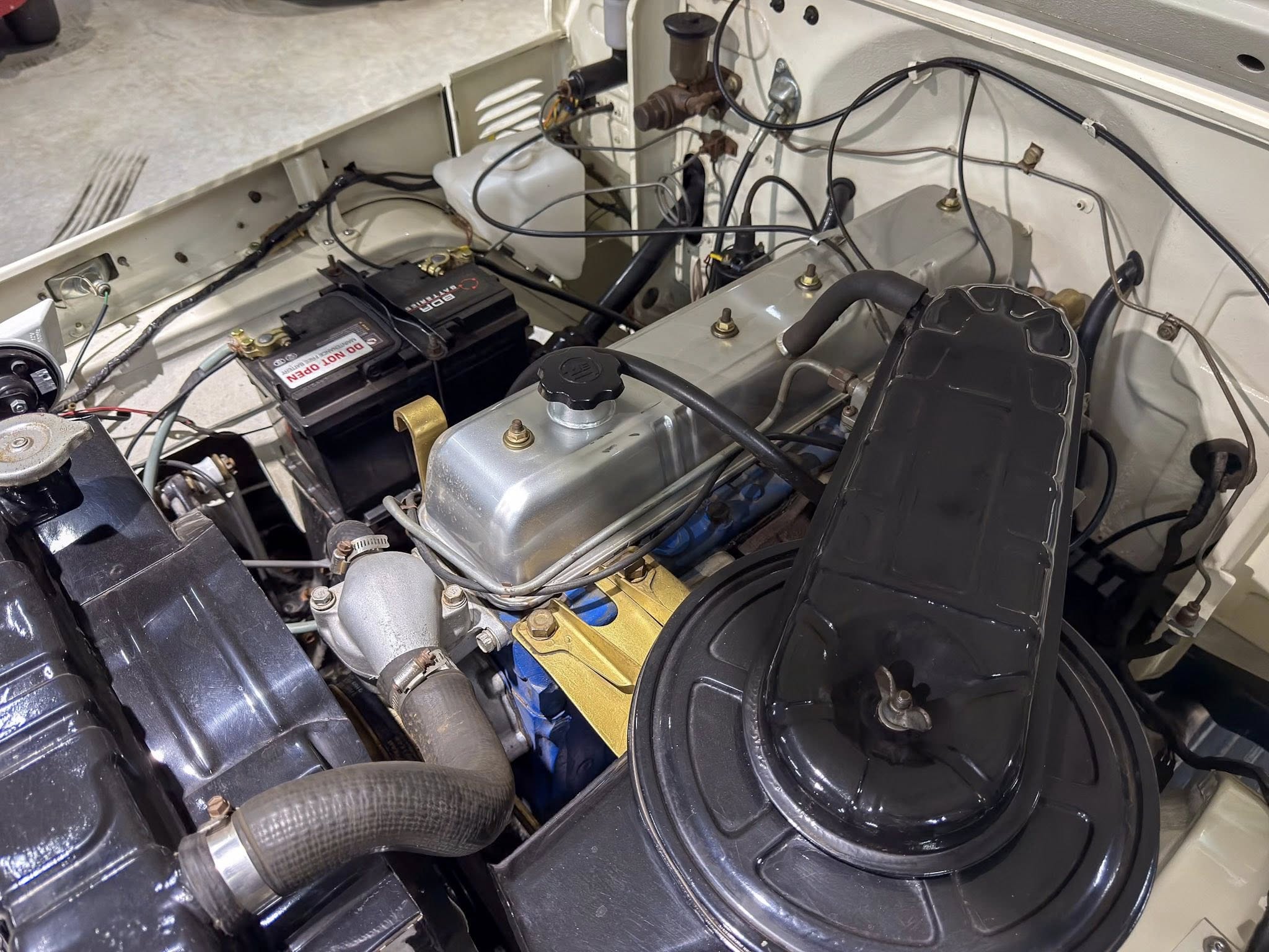 Car engine bay with a battery, hoses, and various wiring; visible components include a coolant reservoir and brake fluid area, wires routed across the firewall, and a black plastic air intake housing in the foreground.